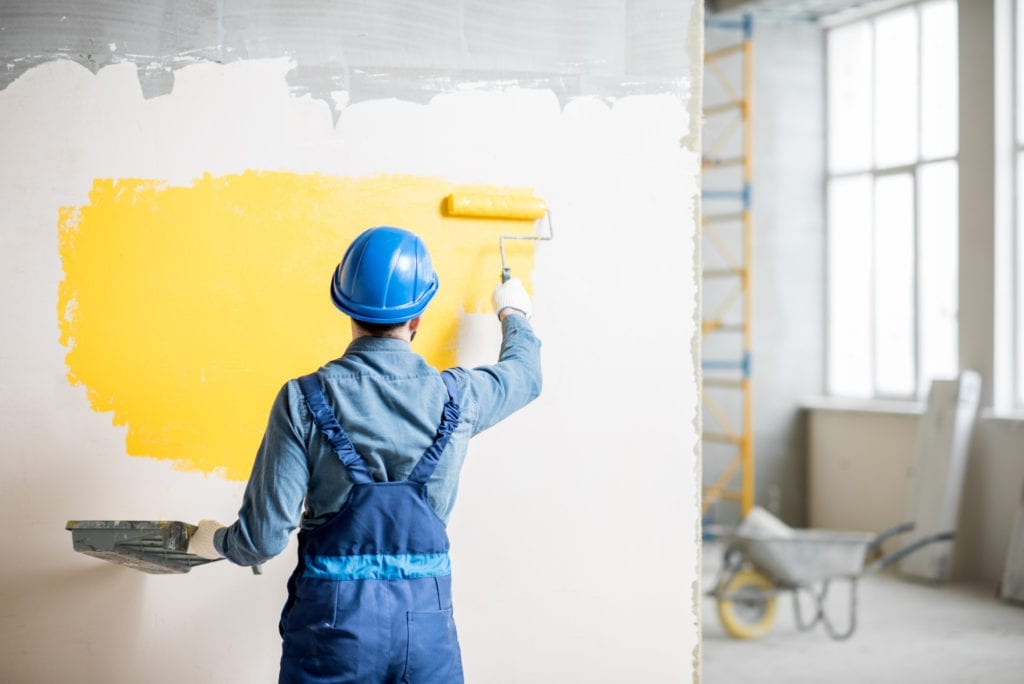 Workman in uniform painting wall with yellow paint indoors
